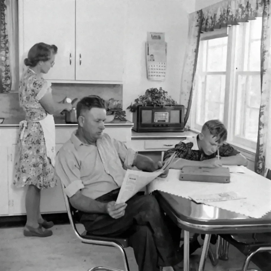 A woman cooks at the stove, a man reads a newspaper at the table, and a boy does homework in a bright 1950s kitchen.