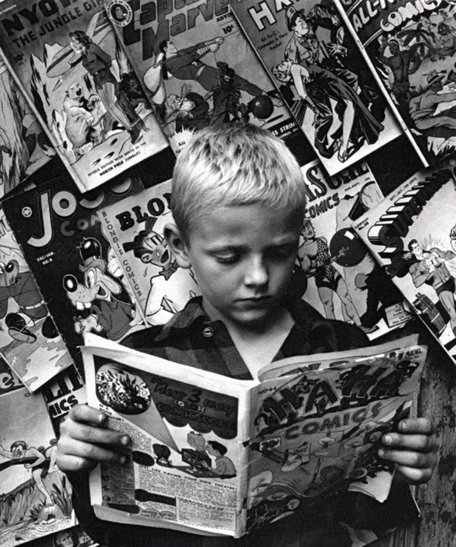 A young boy with short blond hair reads a comic book in front of a wall covered with various colorful comic books, focusing intently on the pages in his hands.