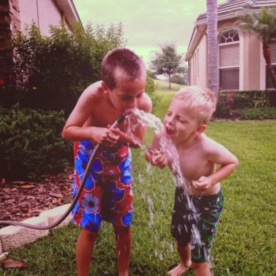 Two young boys in swim trunks drink water from a garden hose on a grassy lawn between houses. One holds the hose while both laugh and enjoy the water on a warm day.