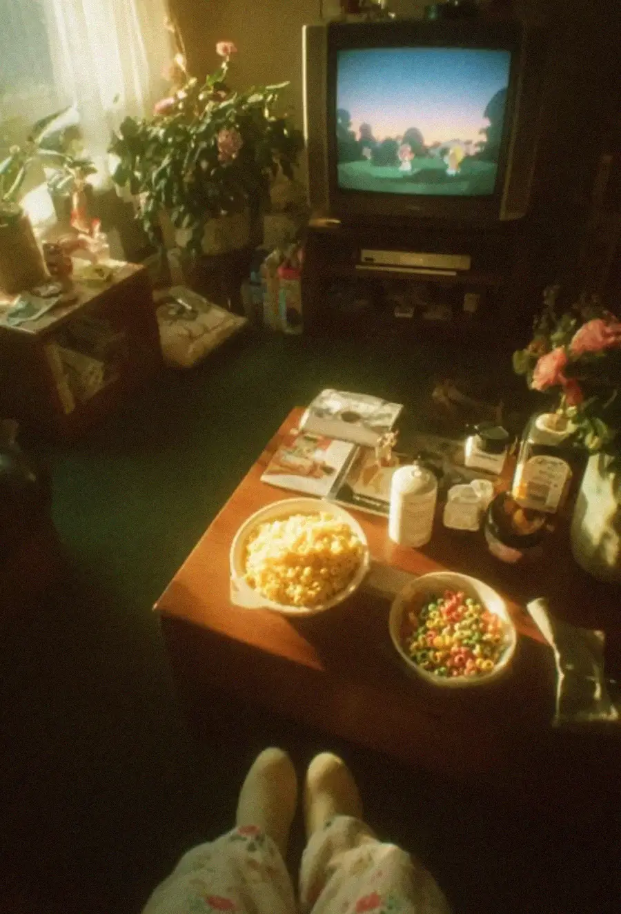A cozy living room with a TV showing a cartoon, a coffee table with cereal, popcorn, syrup, and magazines, and someone’s slippered feet in pajamas visible at the bottom of the image. Sunlight streams through the window.