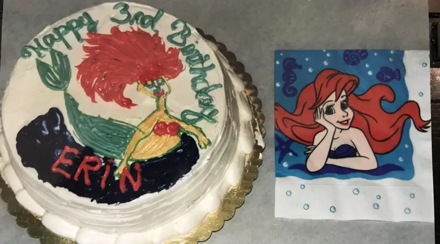 A birthday cake with a poorly drawn mermaid labeled “ERIN” next to a napkin featuring Ariel from The Little Mermaid. The cake says, “Happy 3rd Birthday.”