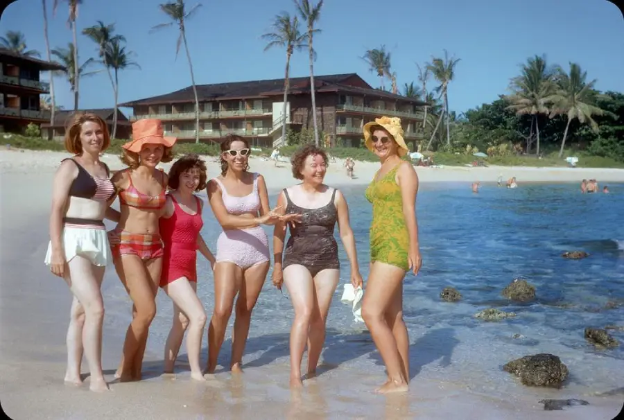 Six women in vintage swimsuits pose and smile on a sandy beach with palm trees, clear blue sky, and beachfront buildings in the background. Some people are swimming in the water nearby.