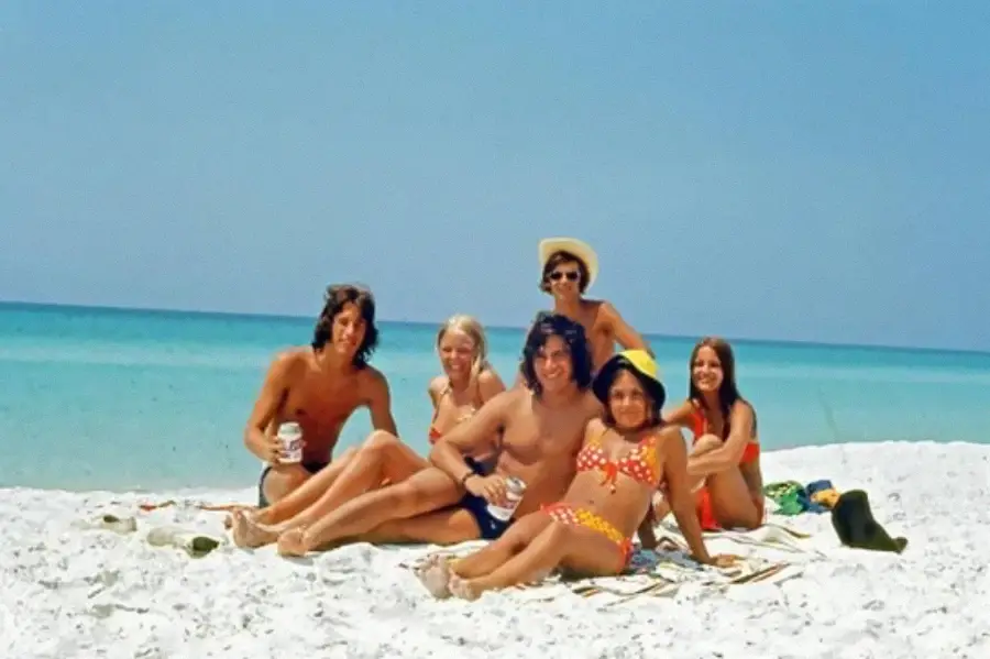 Six young adults in swimsuits sit on a sandy beach near turquoise water, smiling at the camera. The sky is clear and sunny, creating a relaxed, summery atmosphere.