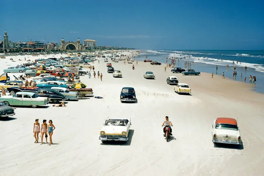 Vintage cars and people on a sandy beach under a clear blue sky; some walk, others drive or relax near the ocean. The scene suggests a lively beach day in a past era.