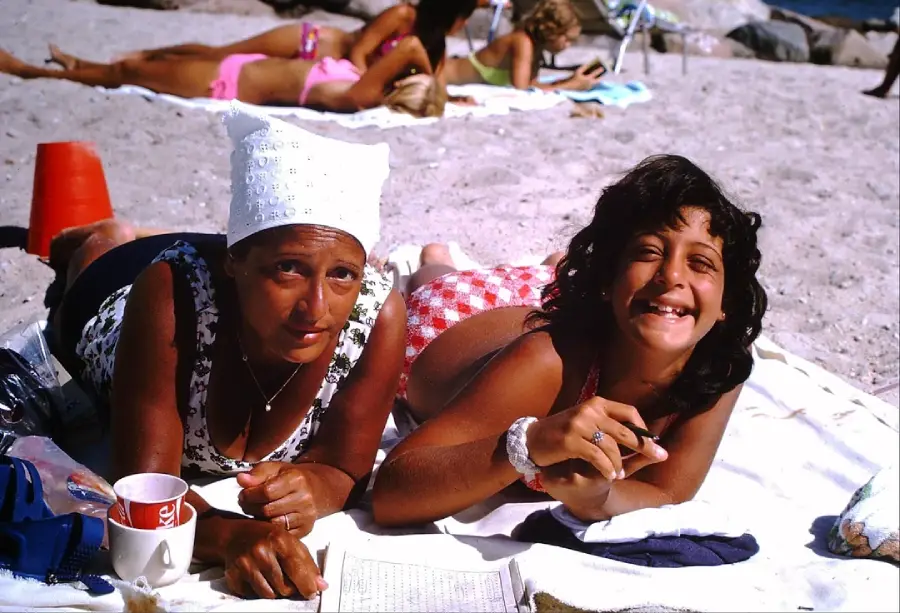 Two people lie on towels at a sandy beach, smiling at the camera. Other beachgoers relax in the background. A red bucket, cup, and paper are nearby. It's a bright, sunny day.