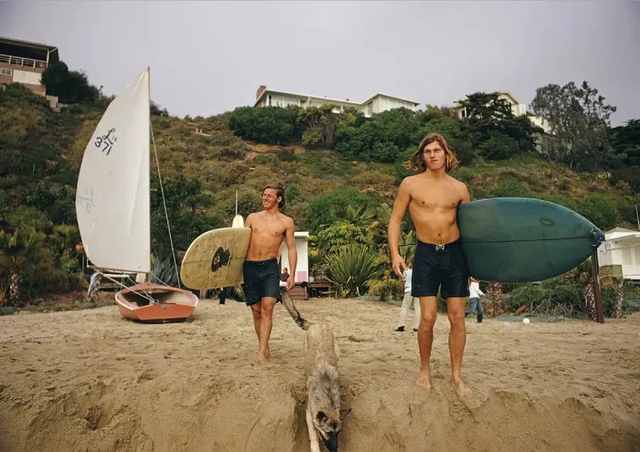 Two men in swim trunks, carrying surfboards, walk barefoot on a sandy beach with a dog. A sailboat and houses on a hill are visible in the background under a cloudy sky.