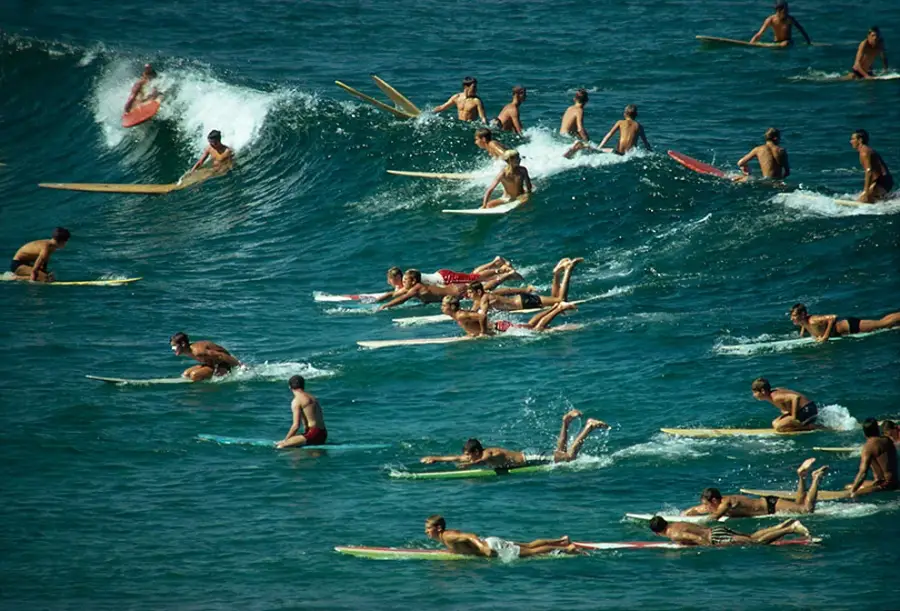 Dozens of surfers paddle on their boards in blue ocean water, with one surfer riding a wave in the background. The scene appears crowded and energetic under clear daylight.