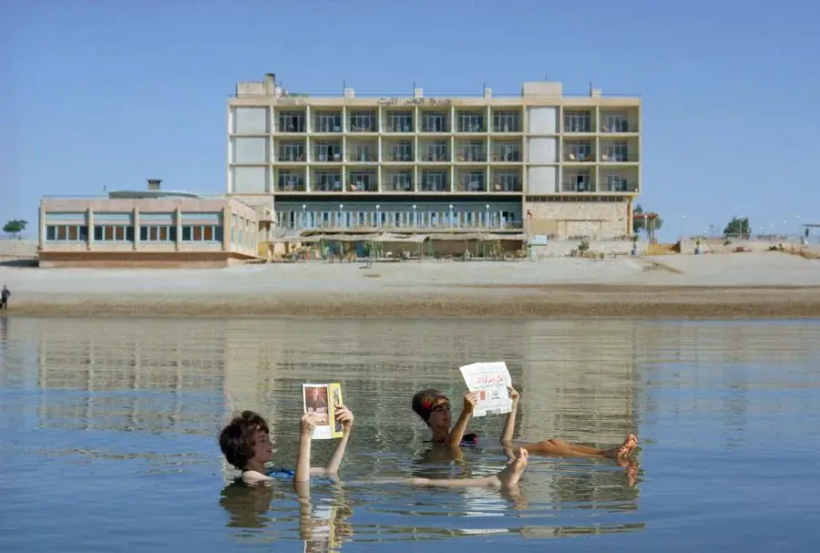 Two people float in water while reading newspapers, with a large, modern hotel building in the background. The sky is clear, and the scene appears calm and relaxed.