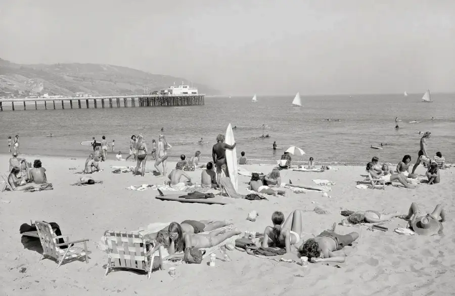 People relax on a sandy beach with towels and chairs, some sunbathing, some standing. Swimmers and sailboats are visible in the ocean. A long pier extends into the water, and hills rise in the background under a hazy sky.