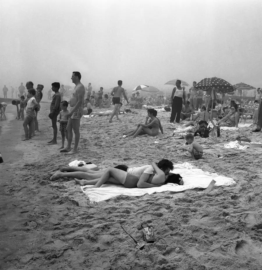 Black and white photo of a crowded beach. People relax on towels, stand, or walk. In the foreground, a couple lies together on a blanket, embracing. Umbrellas and groups are scattered across the sandy shore.