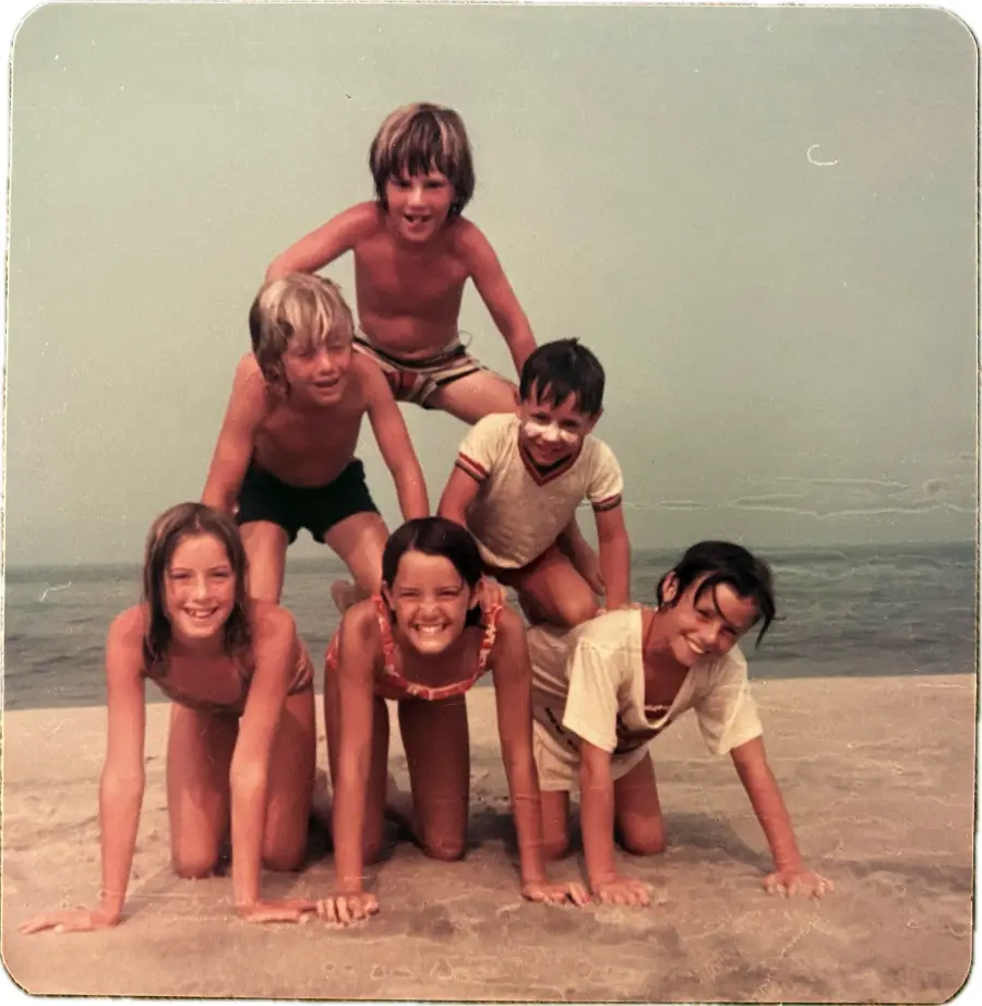 Six smiling children at the beach form a human pyramid, with three crouched on the sand, two kneeling above them, and one boy at the top. The ocean and a hazy sky are in the background.