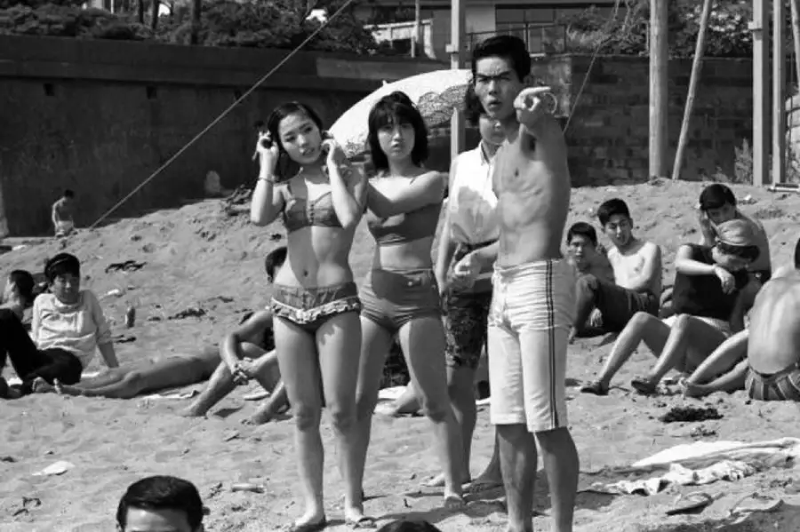 Black and white photo of three people on a busy beach, two women in swimsuits and a man in swim trunks, with the man pointing towards something. Others relax on the sand in the background.