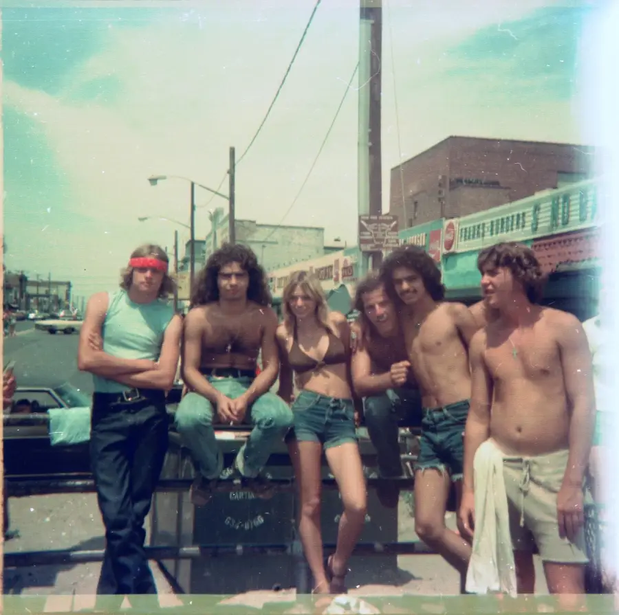 Six young adults in casual, summer clothing pose together on a street in a vintage photo. Three are shirtless, and one wears a red headband. Buildings, cars, and a clear sky are visible in the background.