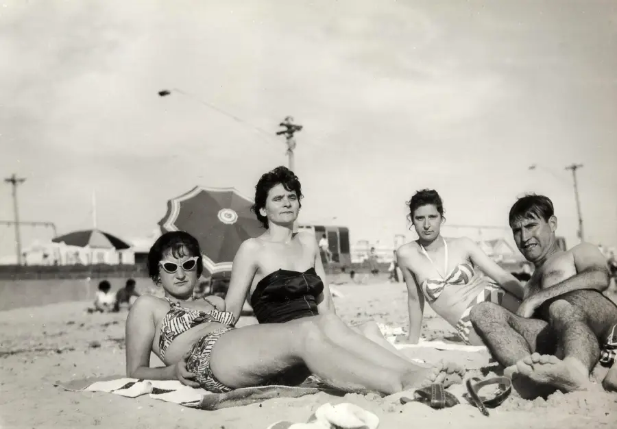Four adults relax on a sandy beach, wearing vintage swimsuits. Two women and two men recline close together, with umbrellas and distant beachgoers visible in the background under a clear sky.