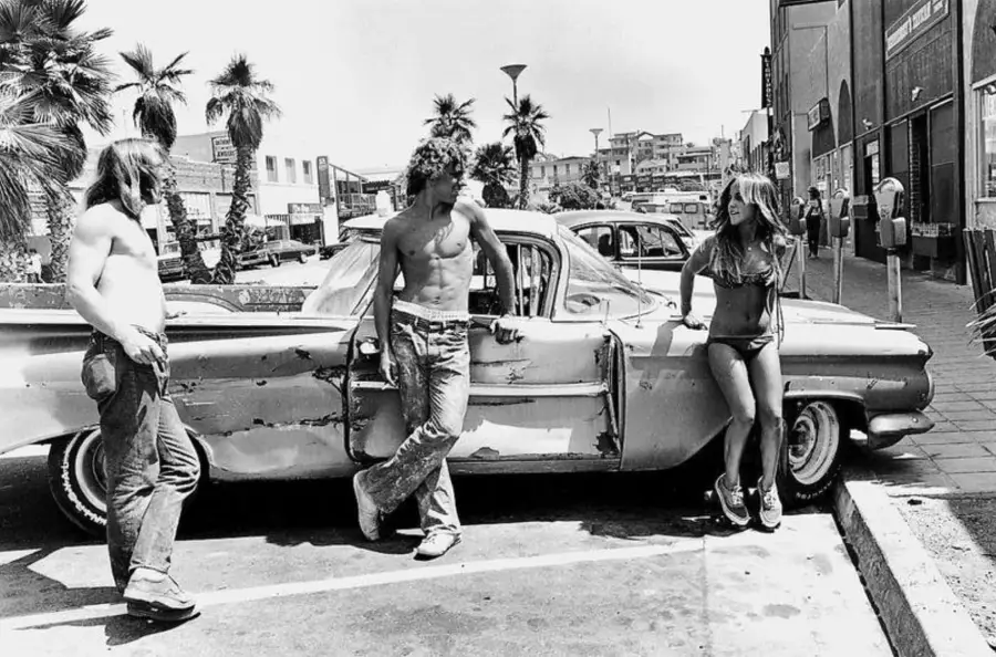 Three young people—two shirtless men and a woman in a bikini—lean against a worn, classic car in a sunny, palm-lined urban street, talking and laughing in a relaxed, casual scene.