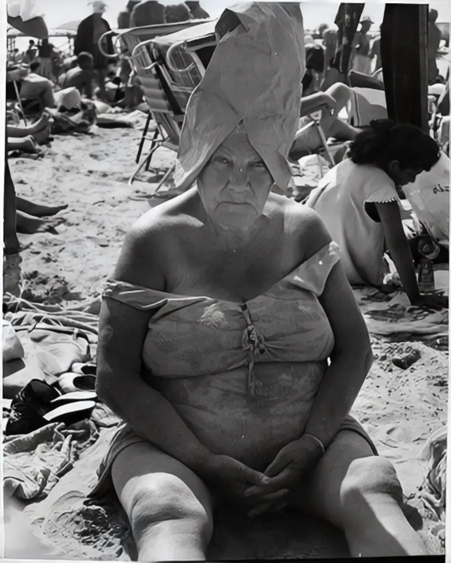 An older woman sits on a crowded beach wearing a swimsuit and a paper bag on her head. She looks directly at the camera, surrounded by other beachgoers and beach chairs.