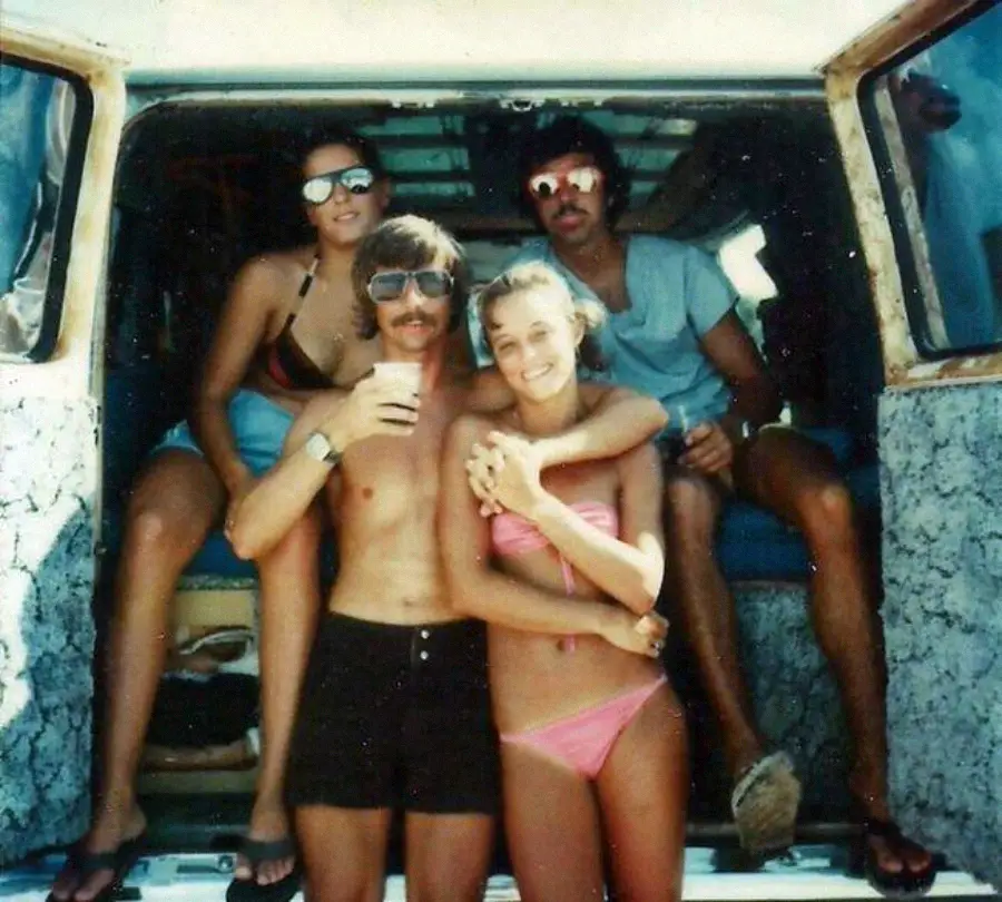 Four people posing in the back of a van; two women in swimsuits and two men in shorts, all wearing sunglasses. The group looks relaxed, enjoying a sunny day together. The van doors are open, revealing a casual, beachy scene.