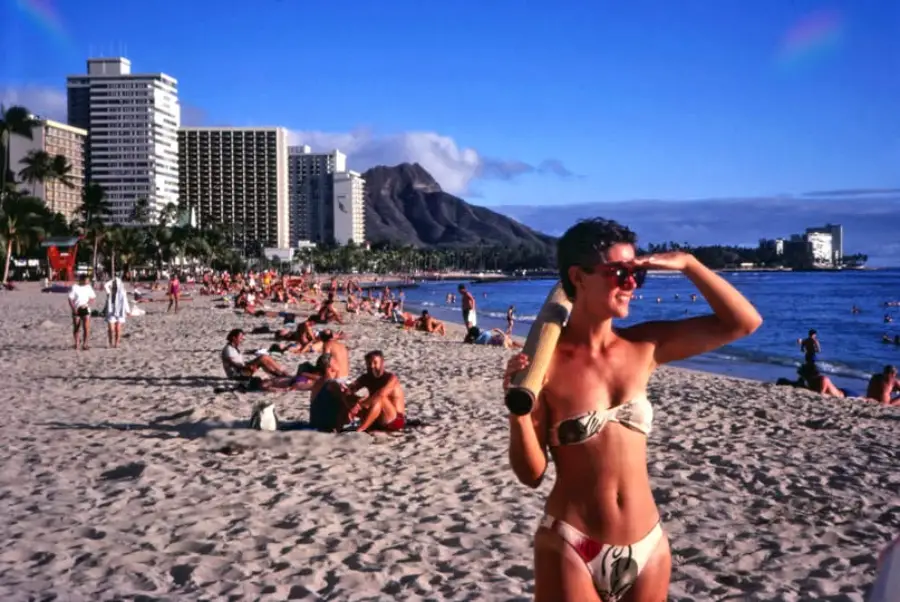 A woman in a bikini stands on a sandy beach with city buildings, people, and a mountain in the background. She holds a towel and shades her eyes, enjoying a sunny day.