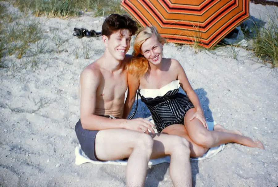 A young man and woman in swimsuits sit smiling on a sandy beach, with grass and an orange-striped umbrella in the background.