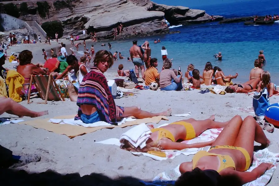 Crowded beach scene with people sunbathing on towels, swimming, and relaxing by the shore. Two people in yellow swimsuits lie face down in the sun. Rocky cliffs and clear blue water are visible in the background.