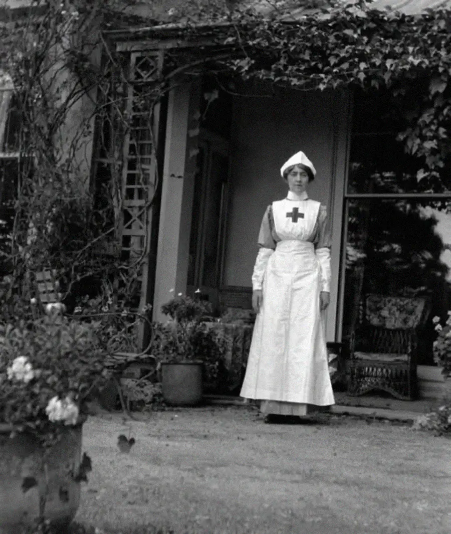 A nurse in a white uniform and cap stands outside a house, near potted plants and flowers. The porch is covered with ivy, and chairs are visible behind her.