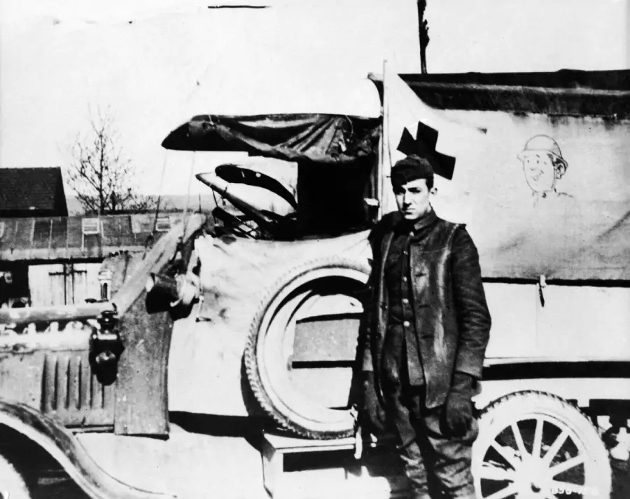 A man in old-fashioned clothing stands next to a vintage ambulance with a Red Cross symbol and cartoon drawing on its side. The vehicle has large spoked wheels and a covered back. The scene appears historical.