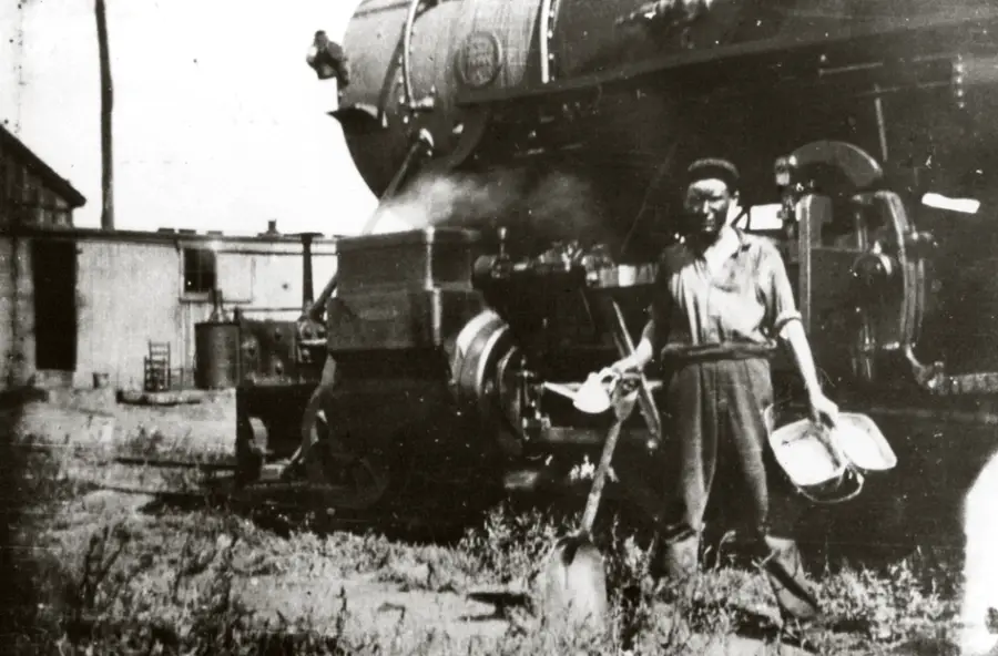 A person stands in front of a steam locomotive holding a shovel and a bucket, wearing work clothes and a cap, appearing to take a break in a train yard with buildings and equipment in the background.