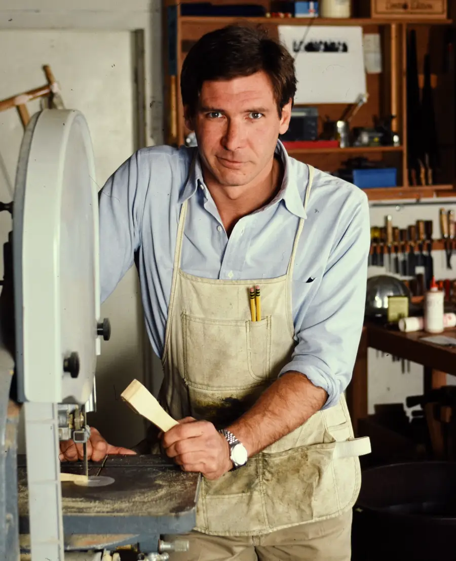 A man wearing a light blue shirt and an apron stands in a woodworking shop, holding a piece of wood by a bandsaw, surrounded by tools and shelves in the background.