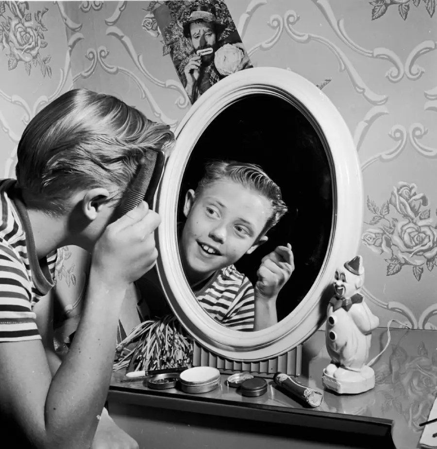 A boy in a striped shirt smiles while combing his hair in front of an oval mirror. Various grooming items and a cartoon rabbit figurine are on the vanity. Vintage wallpaper with floral patterns is in the background.