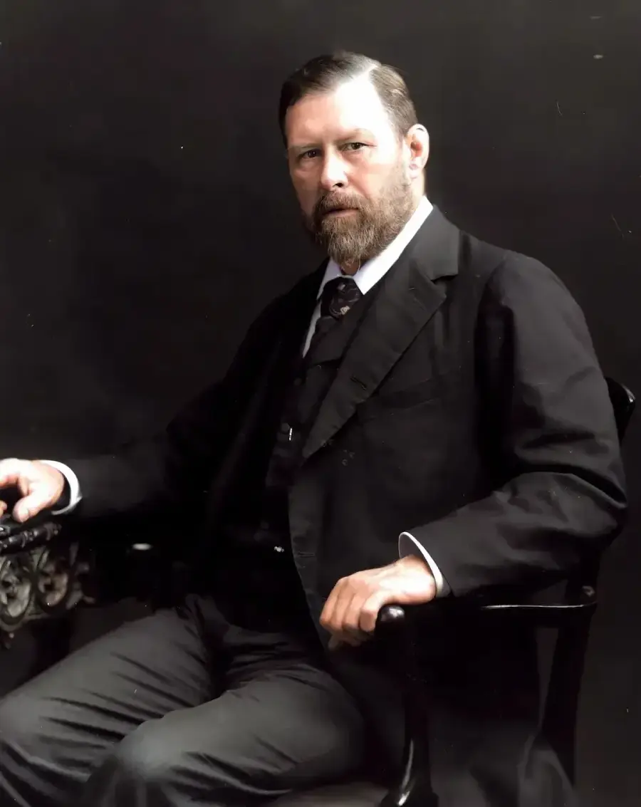 A man with a beard, wearing a dark suit, white shirt, and tie, sits on a black chair against a plain dark background, looking at the camera with a serious expression.