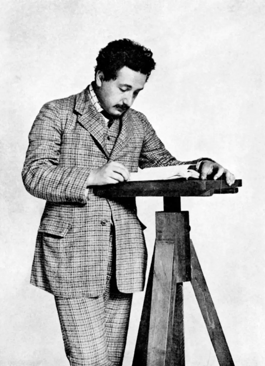 A man in a plaid suit stands at a tall wooden desk, writing in a book while looking down. He has dark curly hair and a mustache. The image is black and white with a plain background.
