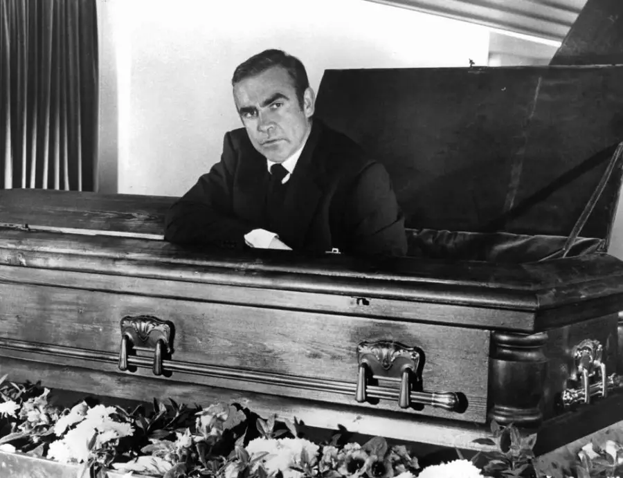 A man in a suit sits upright inside a wooden coffin with the lid open, surrounded by flowers, looking directly at the camera with a serious expression.