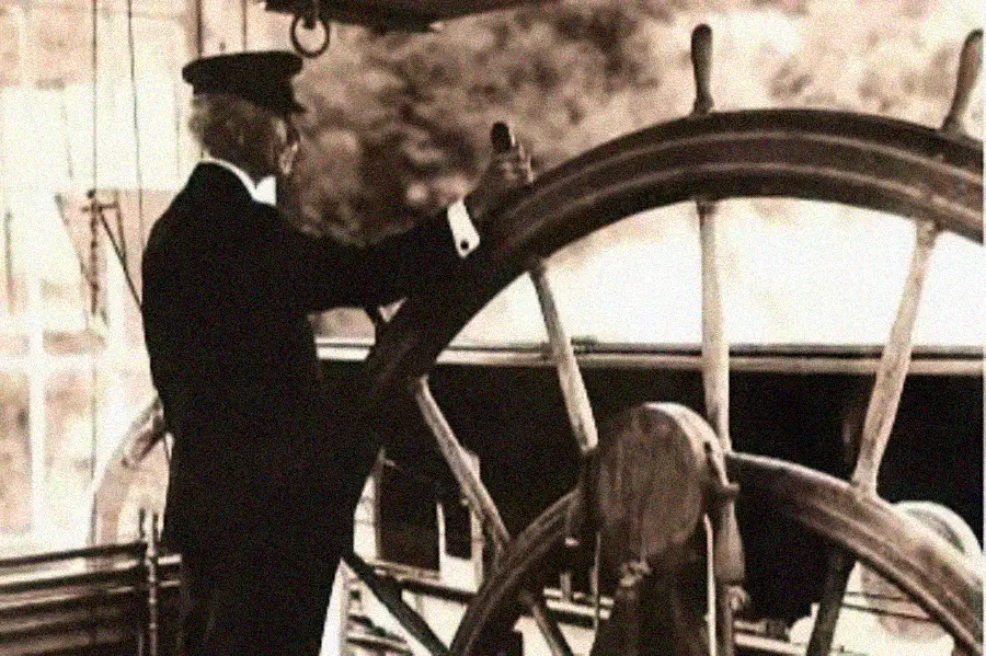 A person in an old-fashioned captain’s uniform stands at the helm, steering a large wooden ship wheel. The image is sepia-toned, giving it a historical feel.