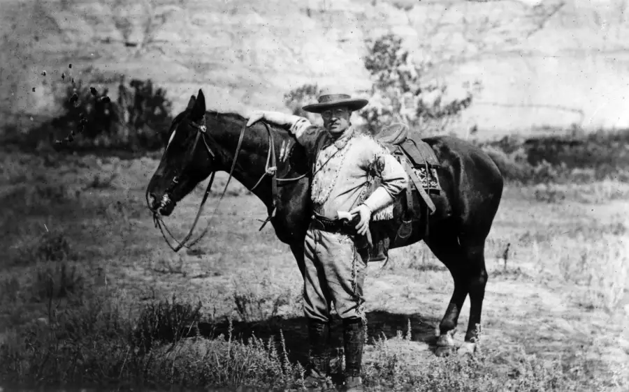 A person in old-fashioned clothing stands outdoors beside a saddled horse, resting one arm on the horse’s back. The background features grass, shrubs, and rocky terrain.