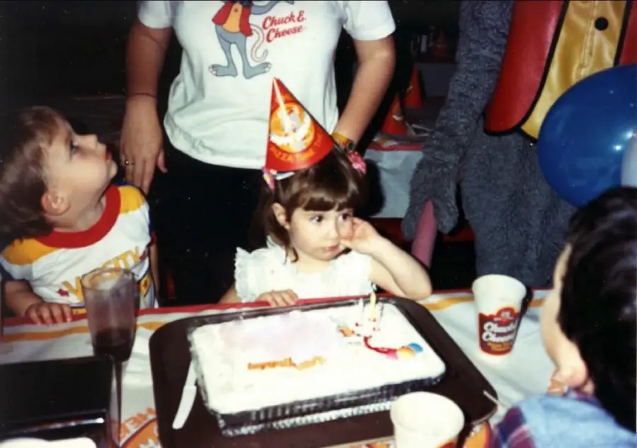 Two children sit at a table with a birthday cake. One child wears a party hat and rests her head on her hand. An adult stands behind them, wearing a Chuck E. Cheese shirt. Party cups and balloons are on the table.