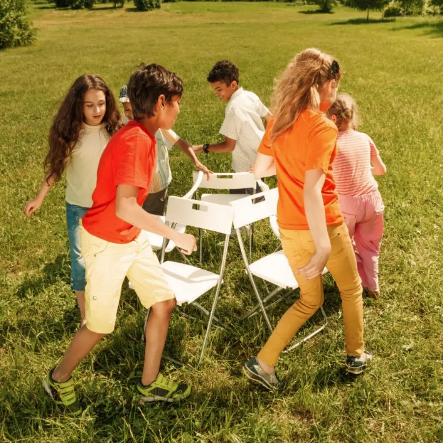 Five children play musical chairs outside on a grassy field, walking around white chairs in a circle. The kids look focused and are dressed in colorful summer clothing. Trees are visible in the background.