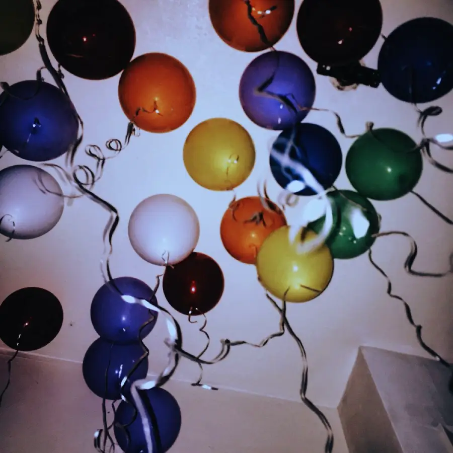 Colorful balloons and curling silver ribbons are attached to a ceiling, creating a festive atmosphere. The balloons are red, orange, yellow, green, blue, purple, and white.