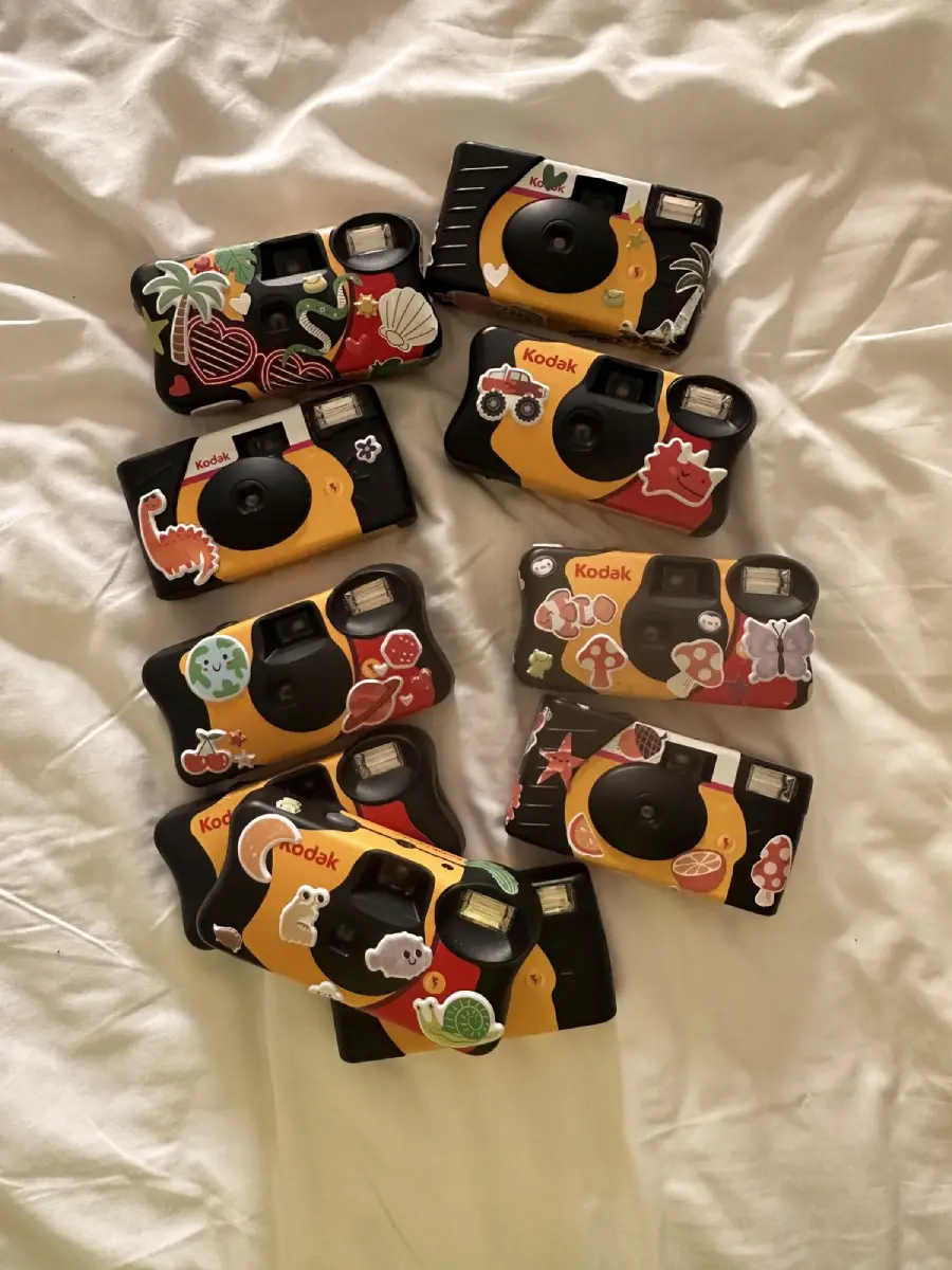 Seven Kodak disposable cameras decorated with colorful stickers are arranged on a white bedsheet. The stickers feature animals, plants, and mushrooms, giving each camera a unique and playful appearance.