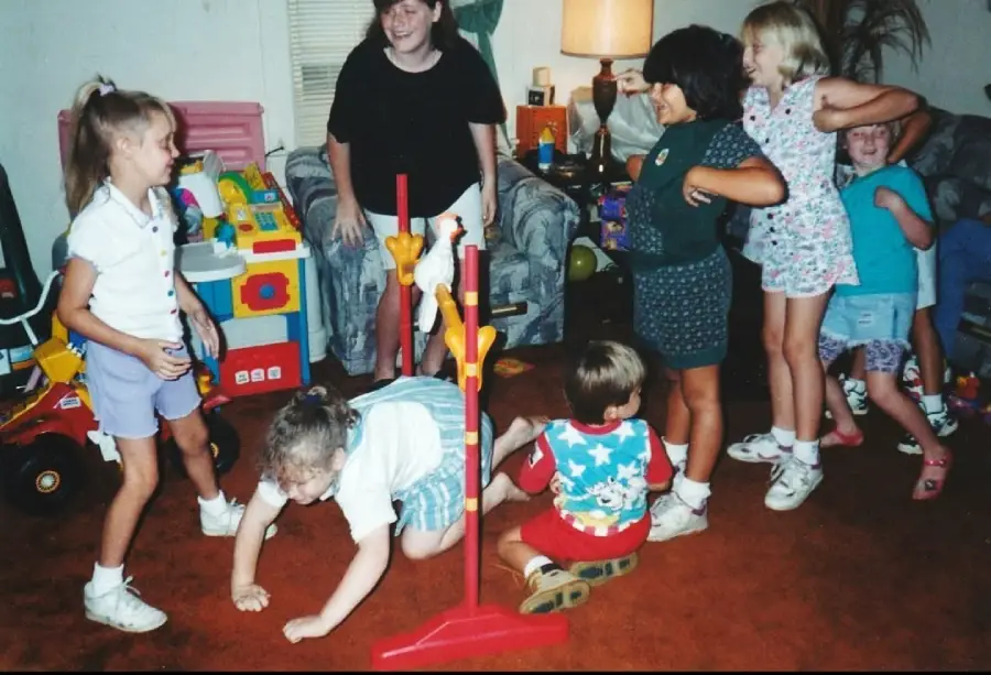 A group of children play limbo indoors, laughing and smiling. One child crawls under the bar while others watch or wait their turn. The room is filled with toys and colorful furniture.