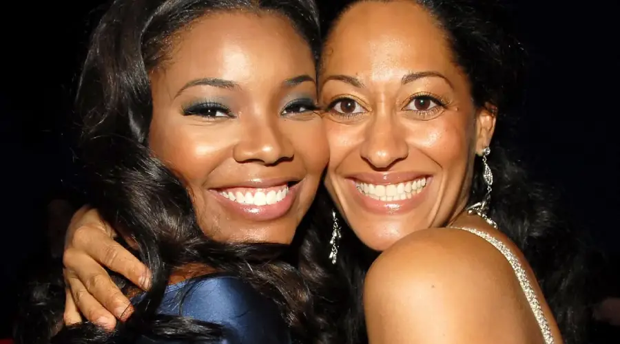 Two women smiling and hugging closely, posing for a photo. Both have long, dark hair and are dressed in evening attire, appearing happy and joyful against a dark background.
