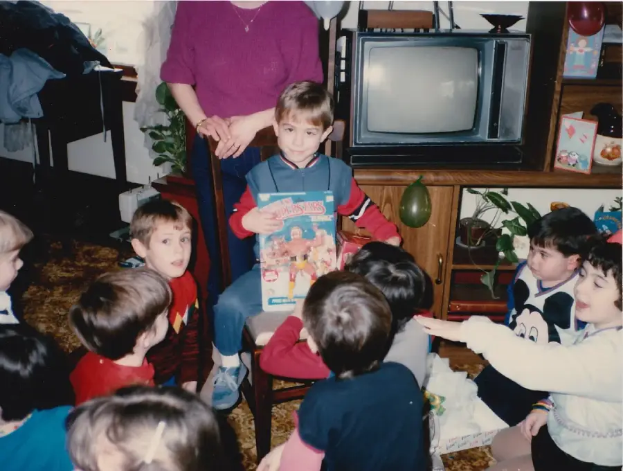 A young boy sits on a chair holding a board game, surrounded by other children at a party. An adult stands behind him. A vintage TV and plants are visible in the background.
