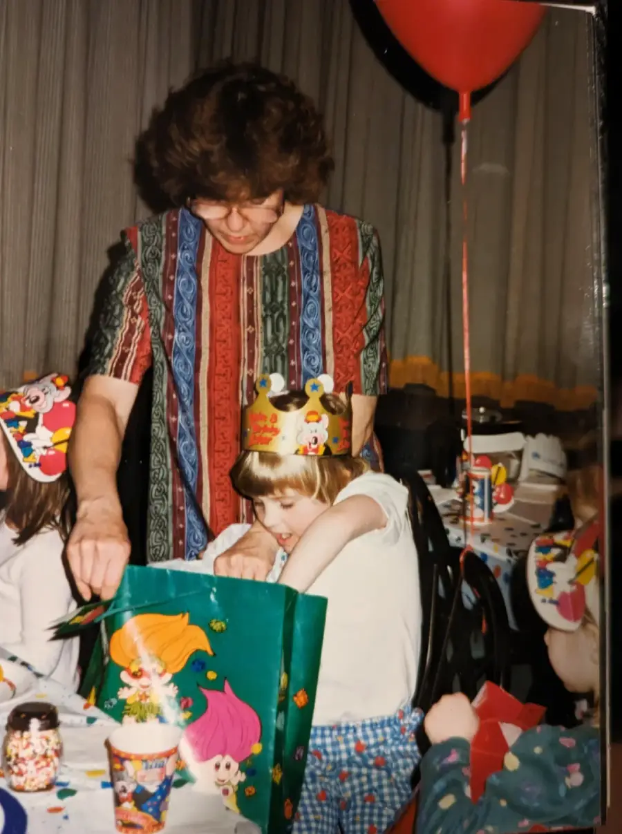 A child wearing a birthday crown reaches into a colorful gift bag while an adult stands behind them, surrounded by other kids at a party table decorated with balloons and cartoon-themed items.