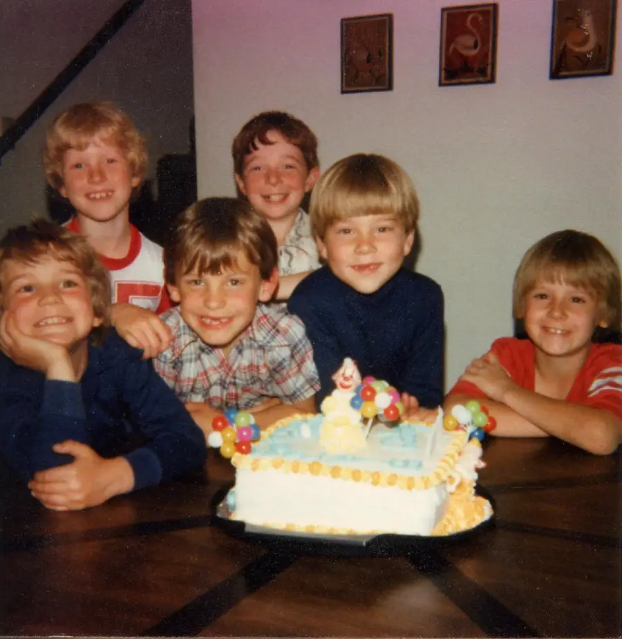 Six smiling young boys gather around a birthday cake decorated with a clown and balloons, sitting at a wooden table in a cheerful indoor setting.
