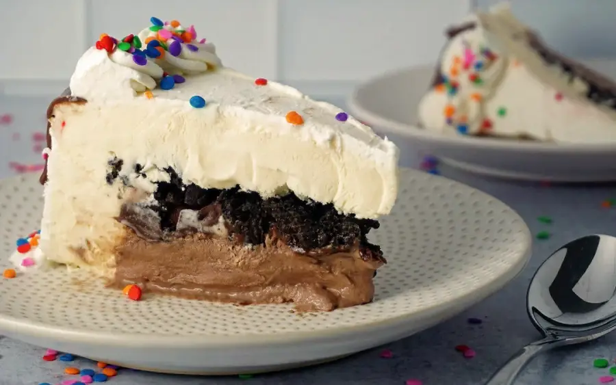 A slice of ice cream cake with chocolate and vanilla layers, topped with whipped cream and colorful sprinkles, sits on a plate next to a spoon. Another slice is in the background.