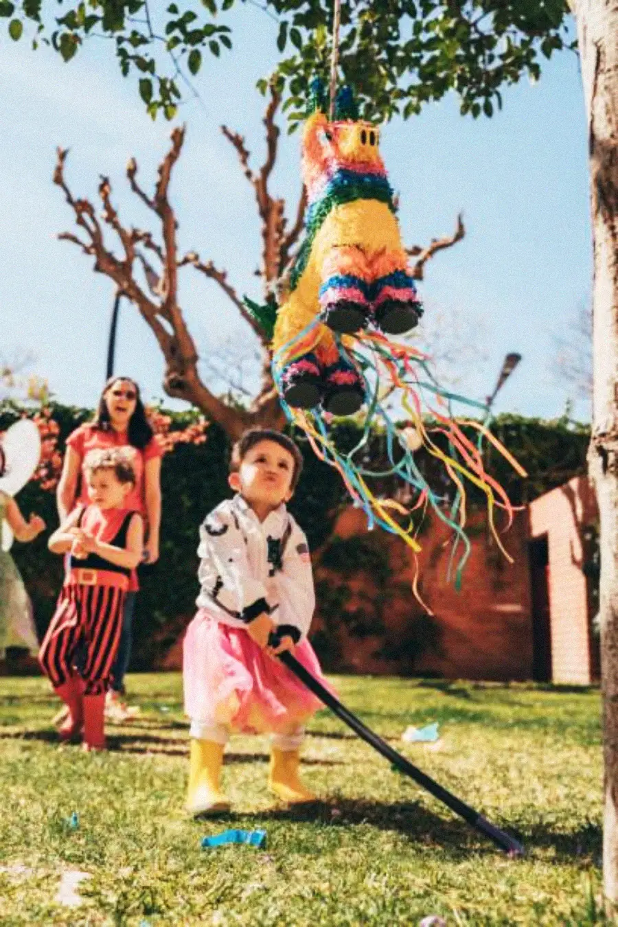 A child in rain boots and costume hits a colorful piñata with a stick at an outdoor party. Other children and an adult watch, surrounded by trees and decorations under a sunny sky.