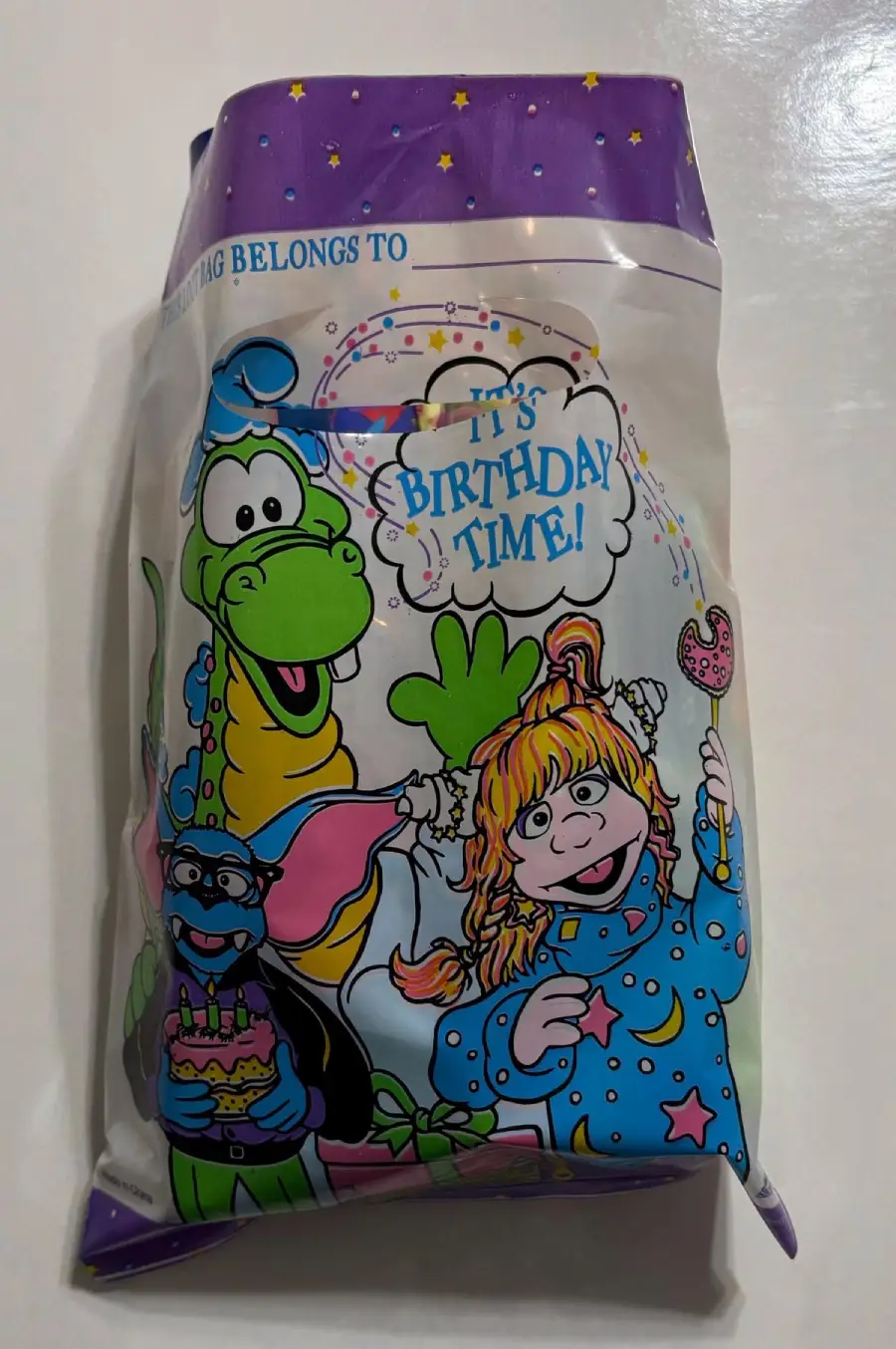 A colorful goodie bag featuring a green dragon, a blue dog with glasses holding a cupcake, and a girl in blue pajamas holding a lollipop, with text that says "IT'S BIRTHDAY TIME!" on a white background.