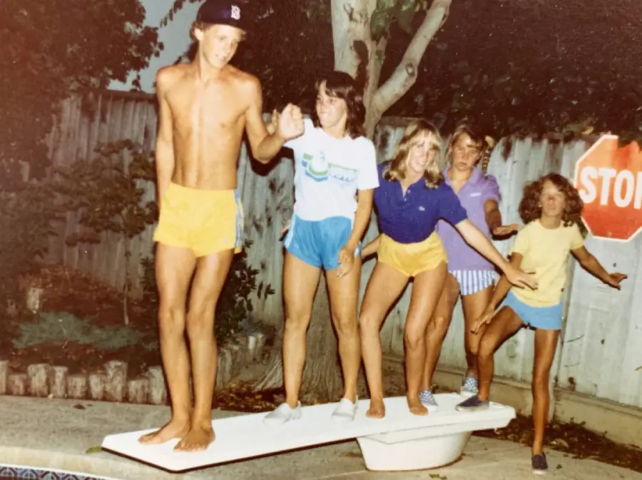 Five kids in summer clothes balance on a diving board by a pool, with trees, a fence, and a stop sign in the background. It appears to be a playful moment in a backyard setting.
