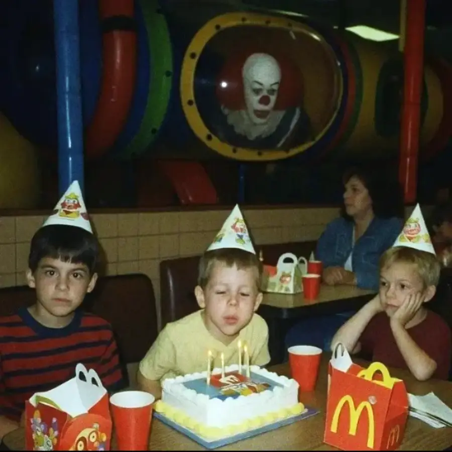 Three boys at a birthday party in McDonald's, wearing party hats. One is blowing out candles on a cake. An adult and Happy Meal boxes are nearby. In the playground tunnel behind them, a person in a clown costume looks on.