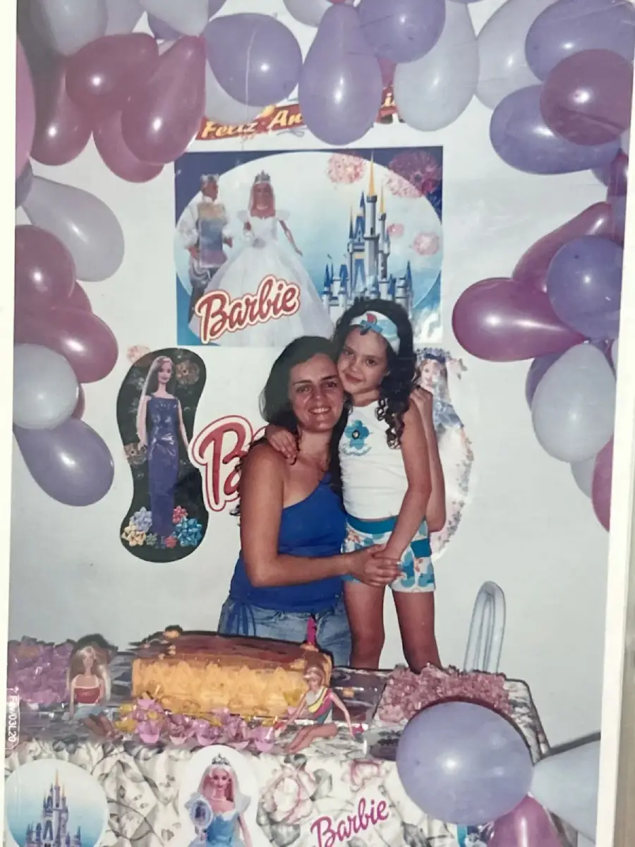 A woman and a young girl smile at a Barbie-themed birthday party with purple and white balloons, posters, and a decorated cake on the table in front of them.
