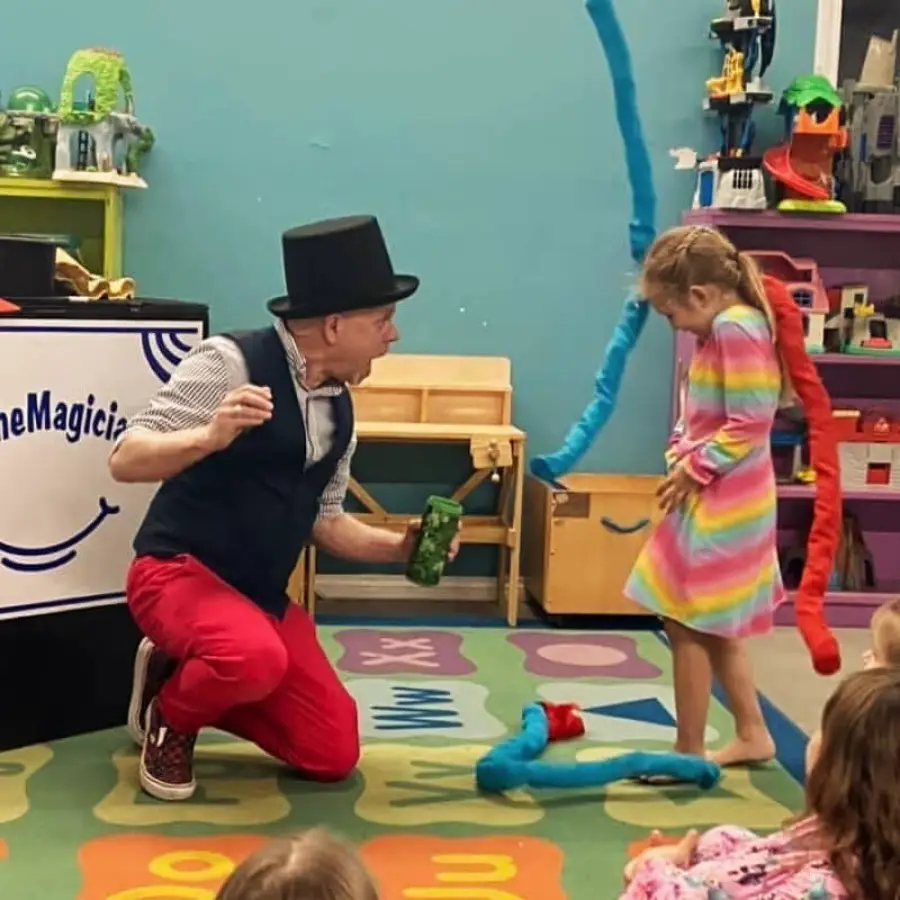 A man in a top hat and red pants performs a magic trick for a young girl in a rainbow dress. Colorful scarves spill from a container as children watch. The setting appears to be a playroom or classroom.